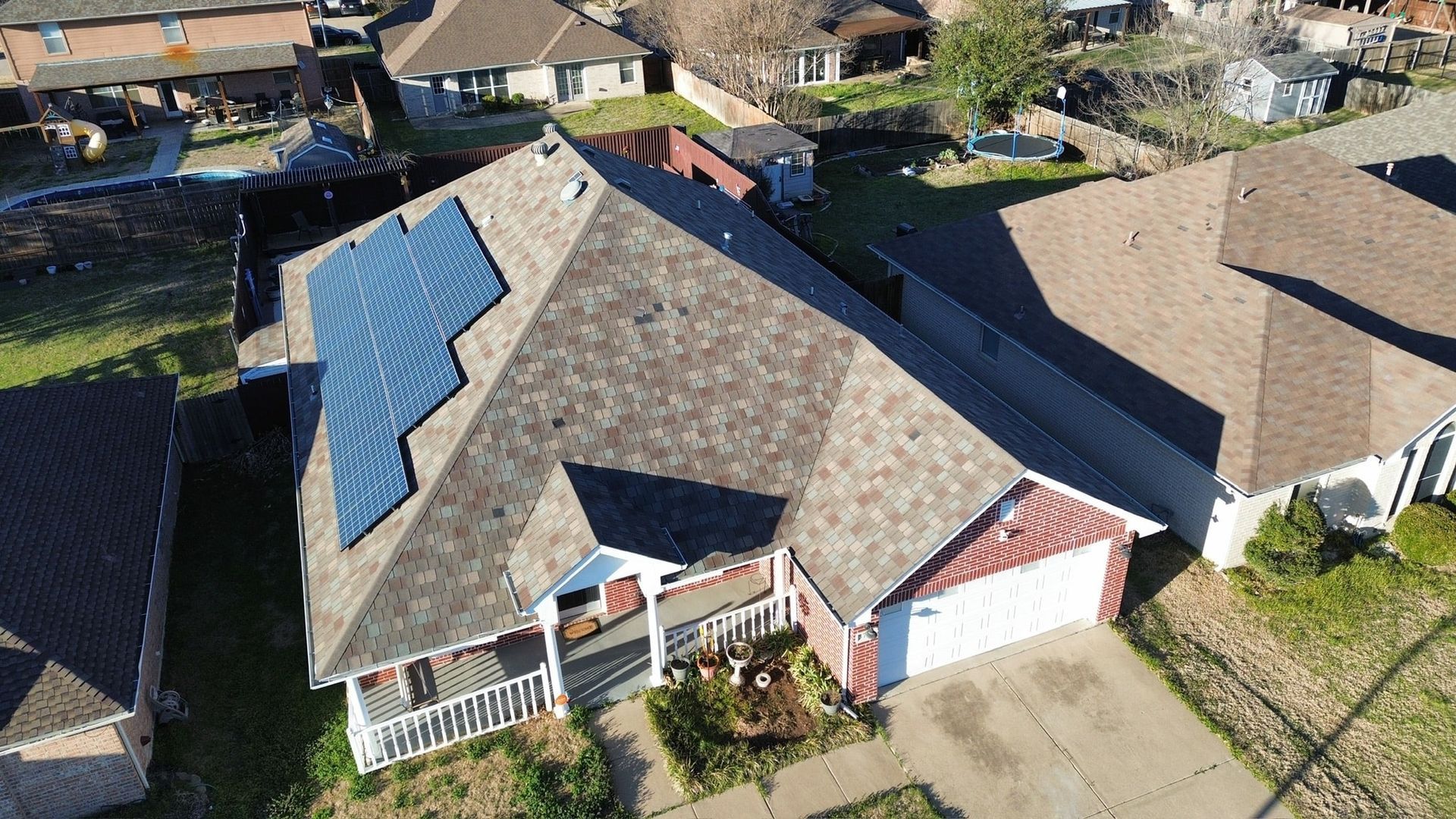 Aerial view of a house with solar panels on its roof, surrounded by other houses and greenery.