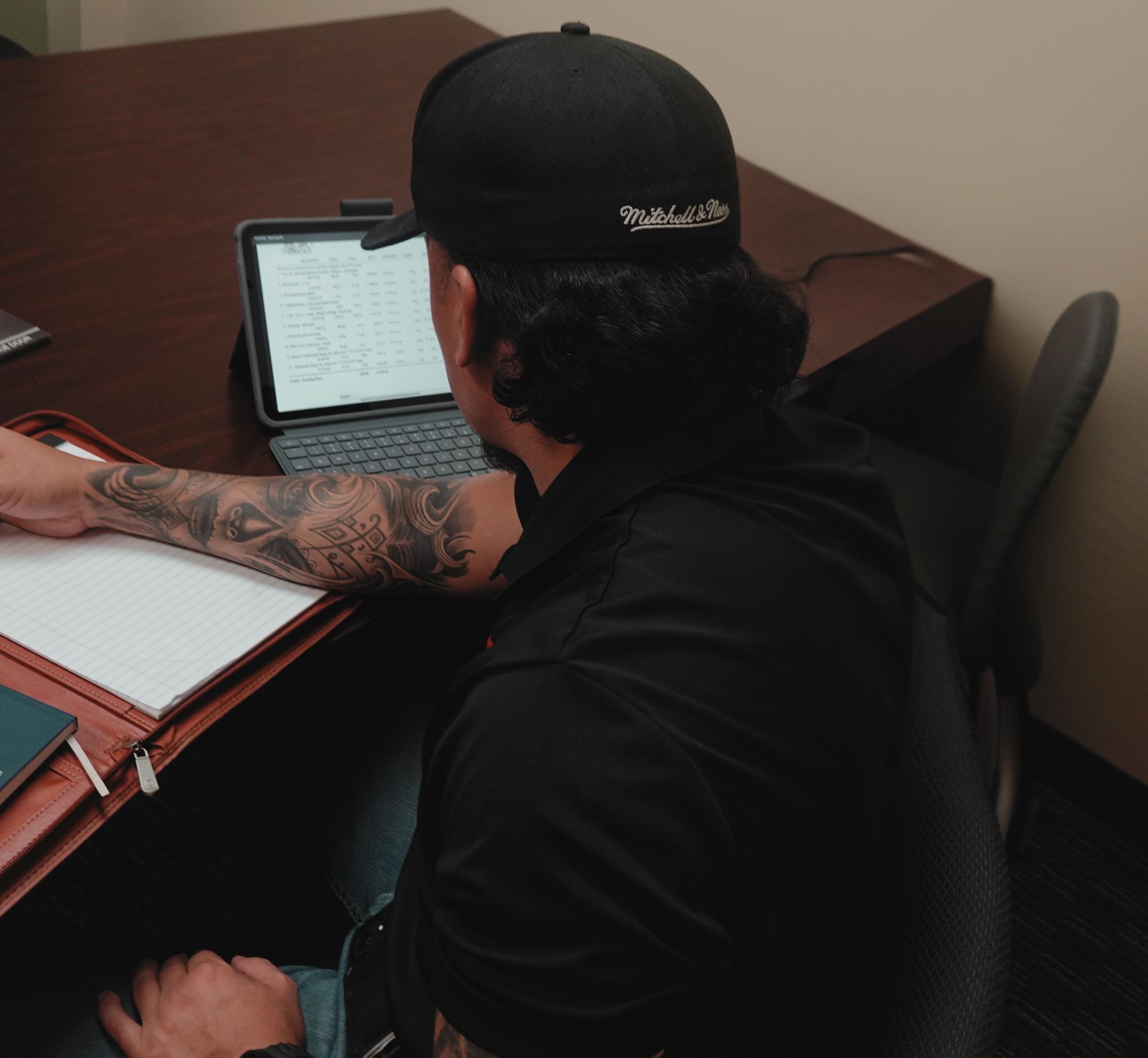 Man with tattoos, black cap, and shirt working at a desk with an iPad, notebook, and pen.