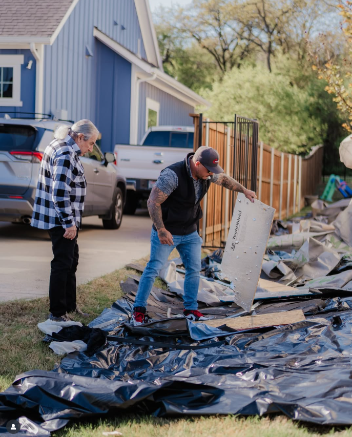 Two people inspecting debris in a yard. Man holds a board; a light blue house is in the background.