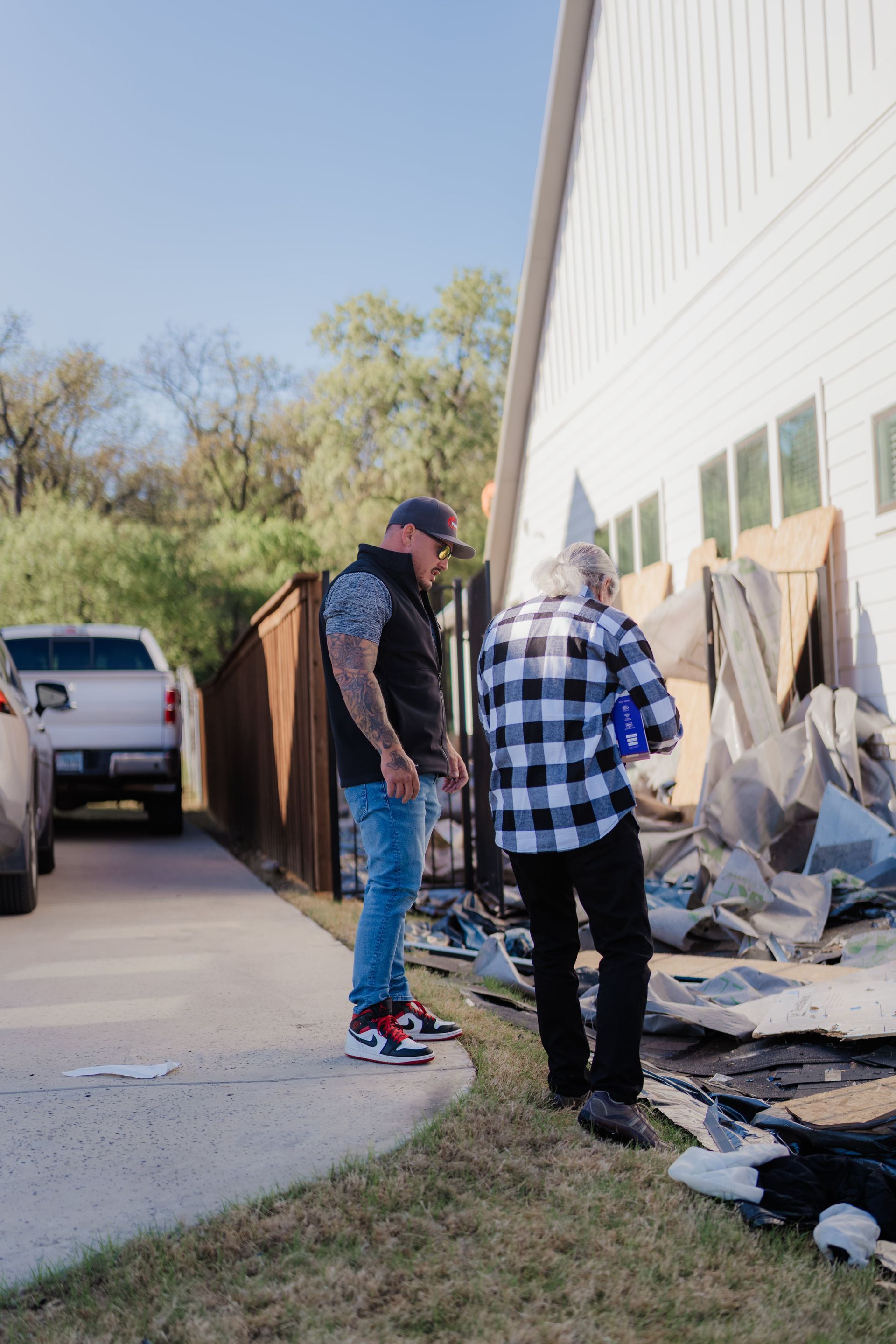 Man and woman surveying damage outside a white house with debris; truck parked in driveway, fence.