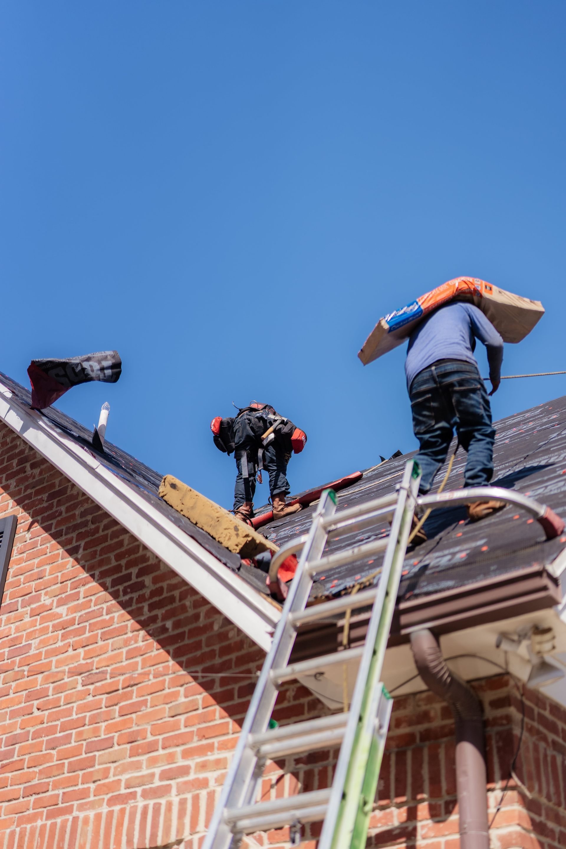 Two roofers on a brick house roof, one carrying a bundle, the other working, blue sky.