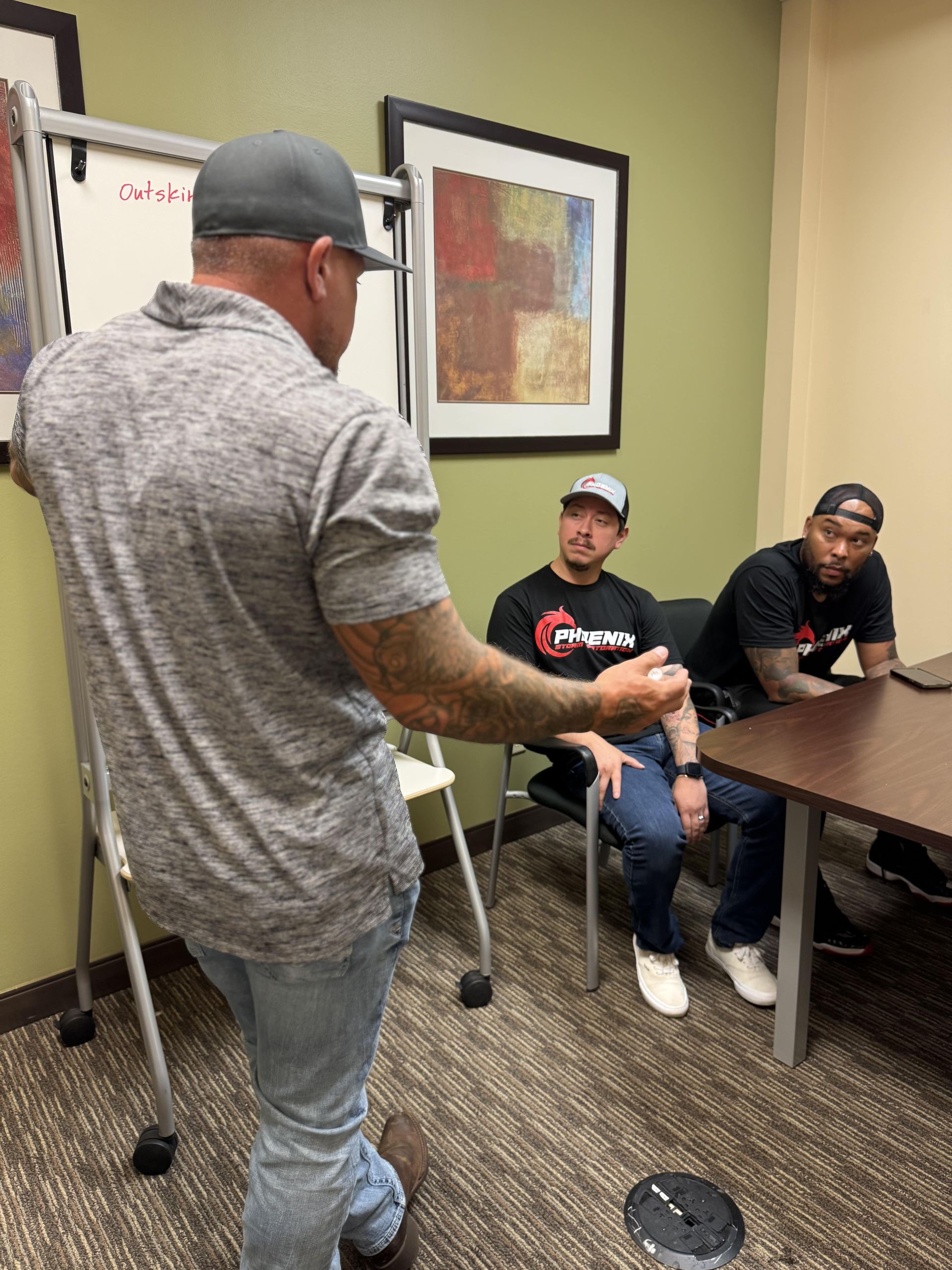 Man in a gray shirt gestures towards a whiteboard, addressing two men in a room.