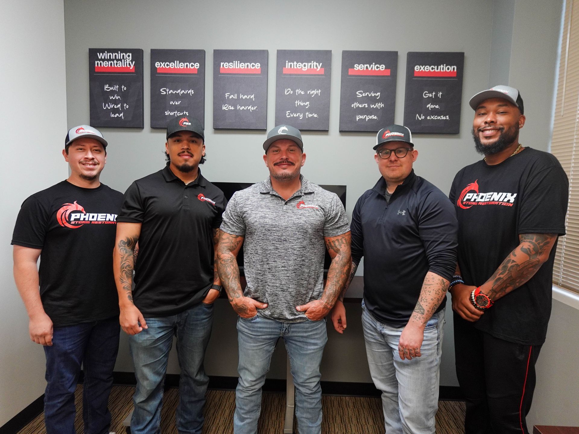 Five men smiling, wearing hats and branded shirts, standing in front of wall art, inside an office.