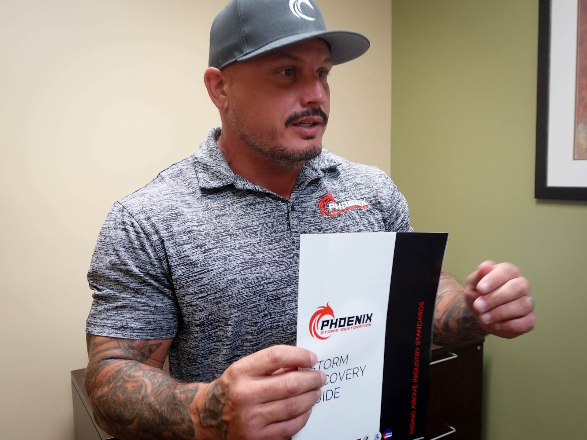 Man in grey cap and shirt holds a document, talking in an office setting.