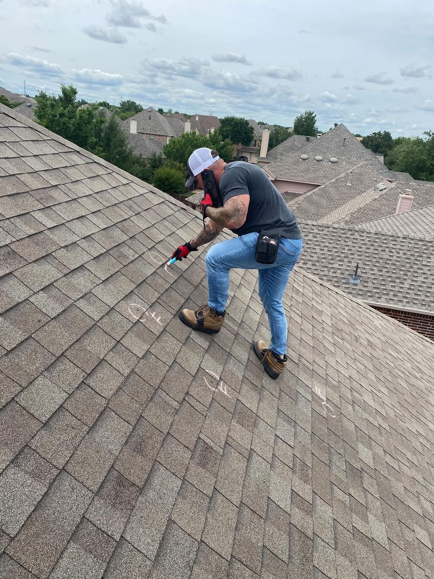 Man on a rooftop with tools, assessing shingles. Wearing safety gear and jeans. Houses in the background.