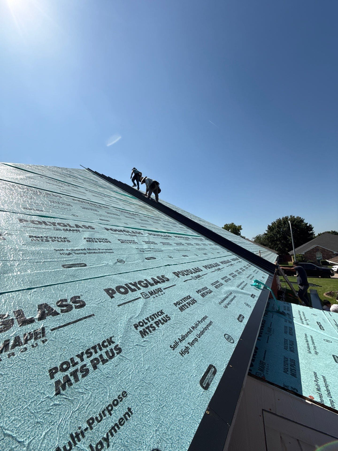 Two roofers installing roofing material on a sunny day. Blue underlayment visible, clear sky.