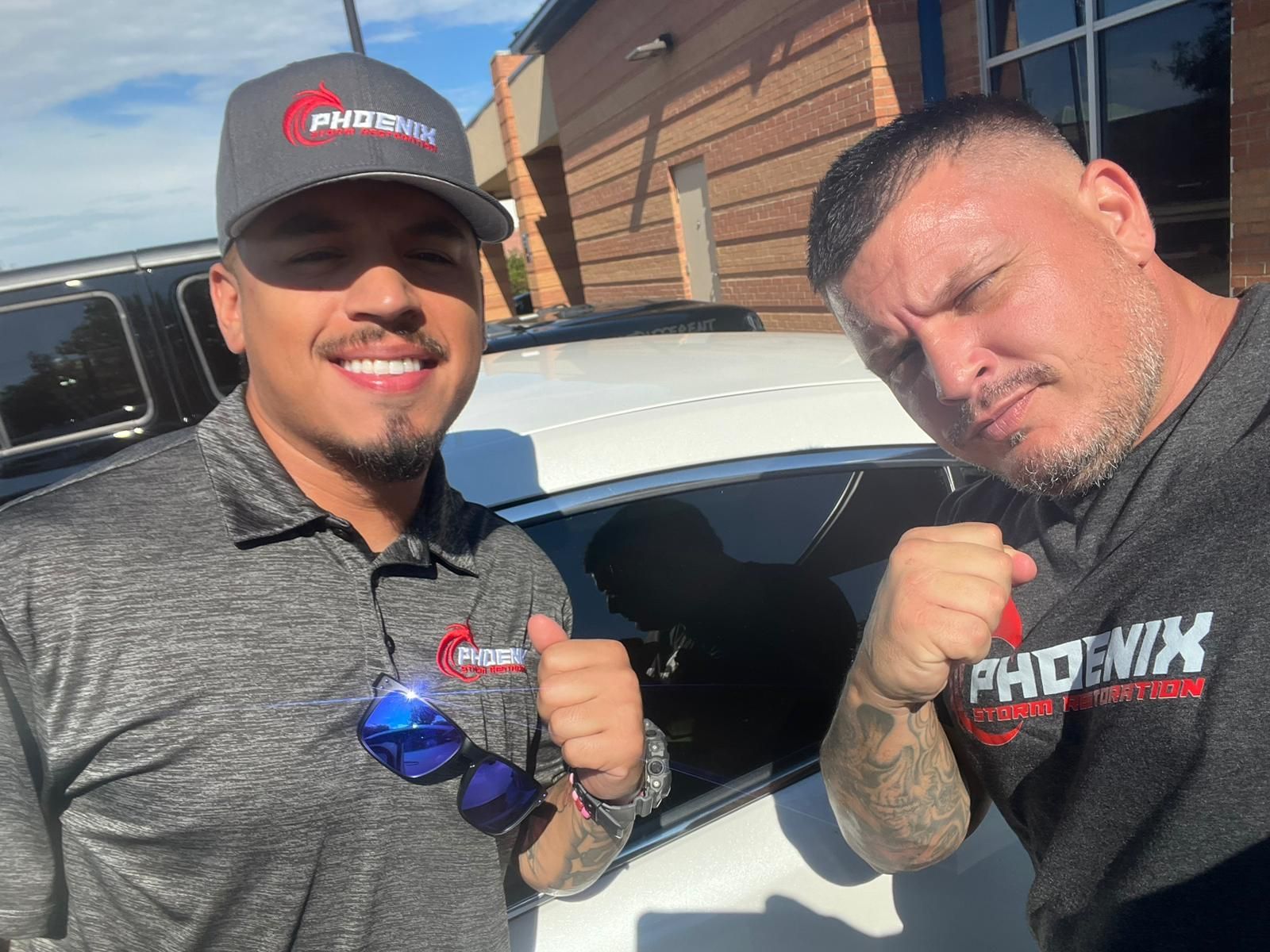 Two men smiling, in gray Phoenix shirts, near a car. One wears a hat.