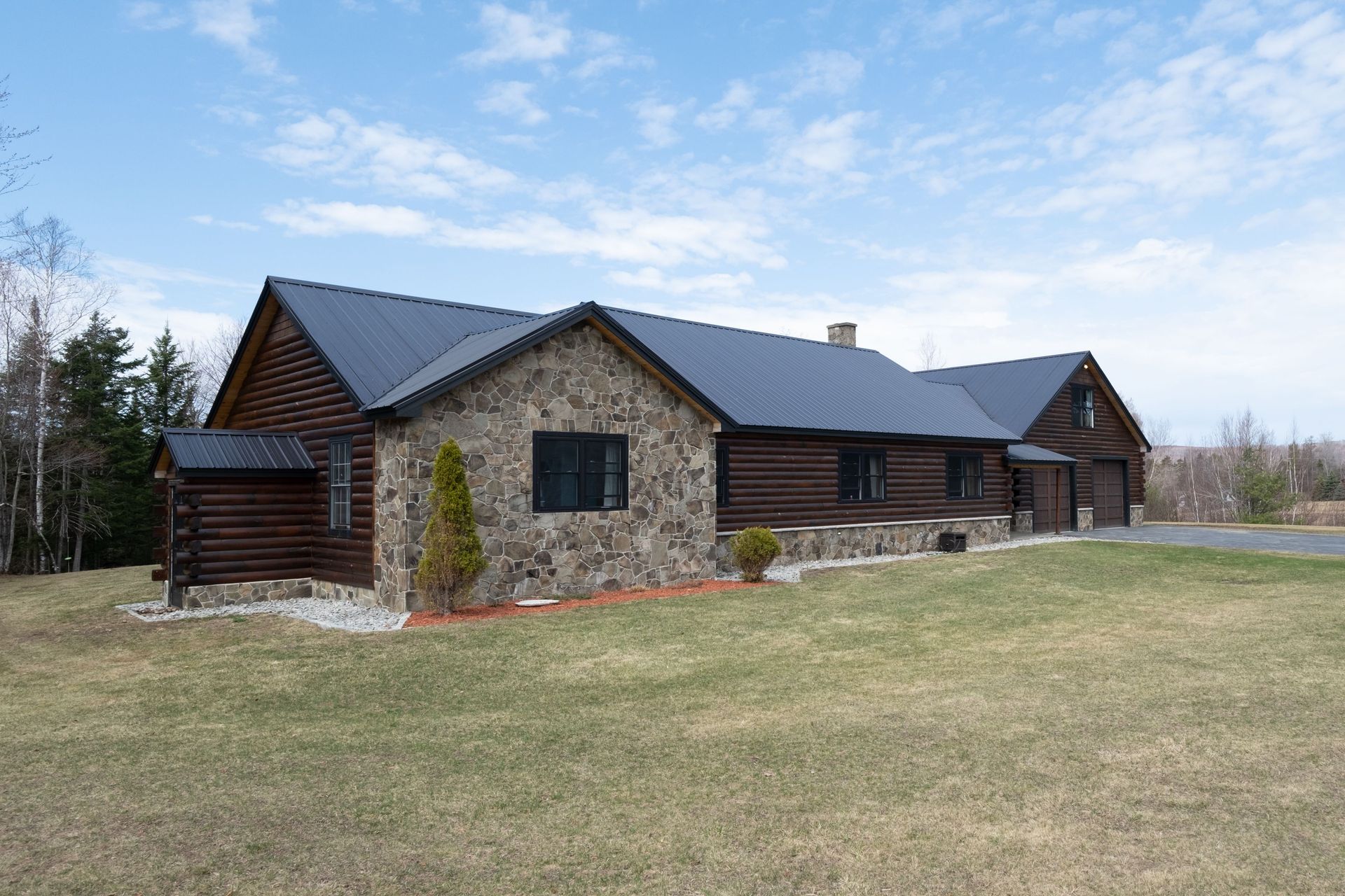 Log cabin with stone facade and dark roof, set on a grassy lot under a cloudy sky.