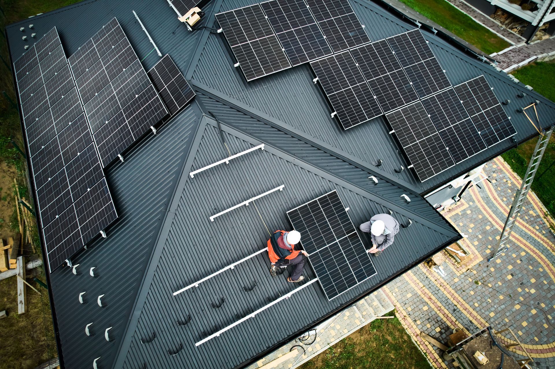 Solar panels are being installed on a dark grey roof by two workers in safety gear.