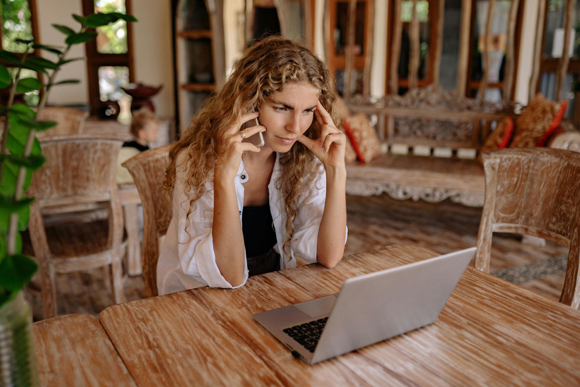 Woman with curly hair on phone, looking stressed at laptop on wooden table.