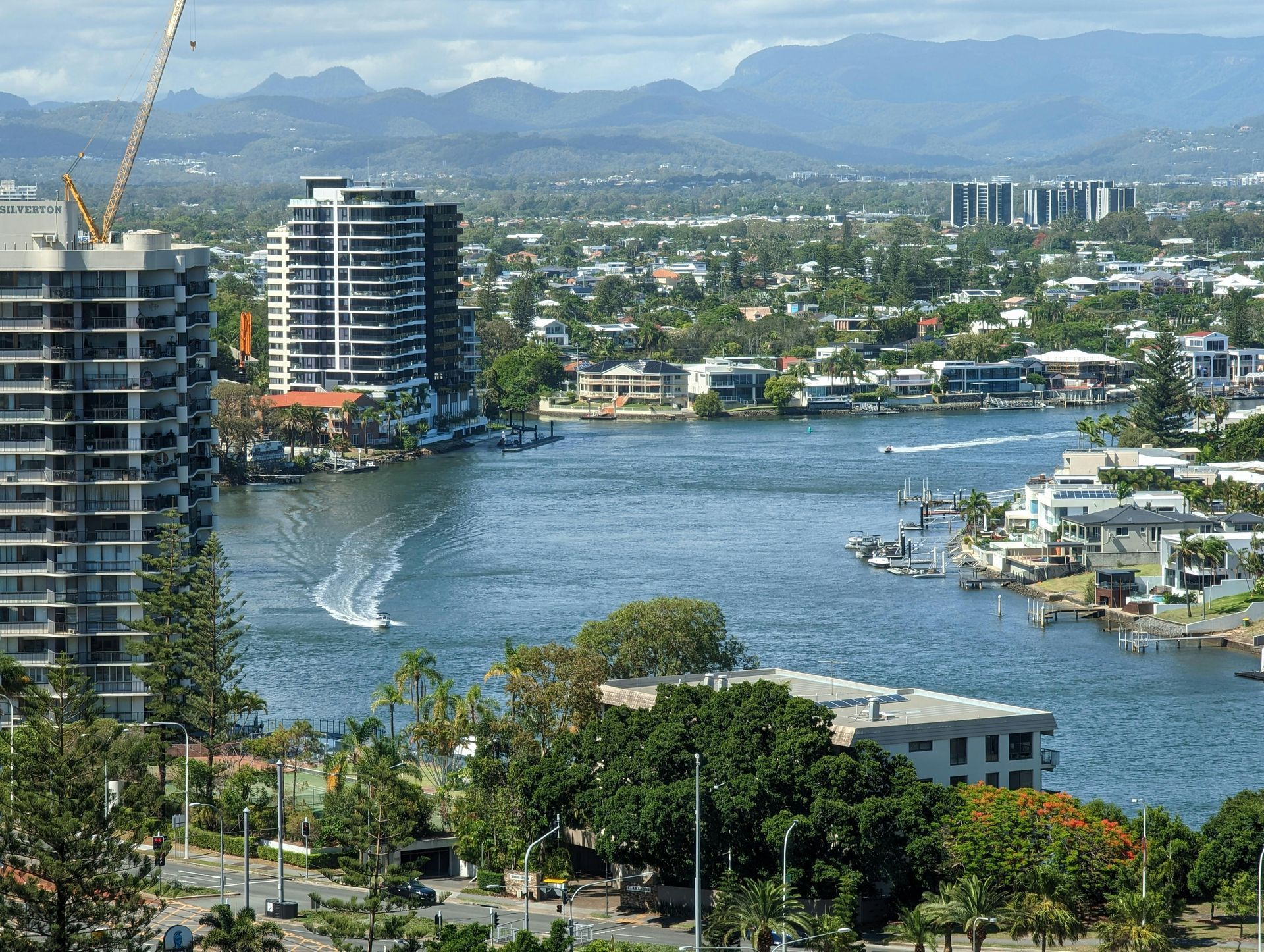 Riverfront cityscape with high-rise buildings, boats, and mountains in the distance under a clear sky