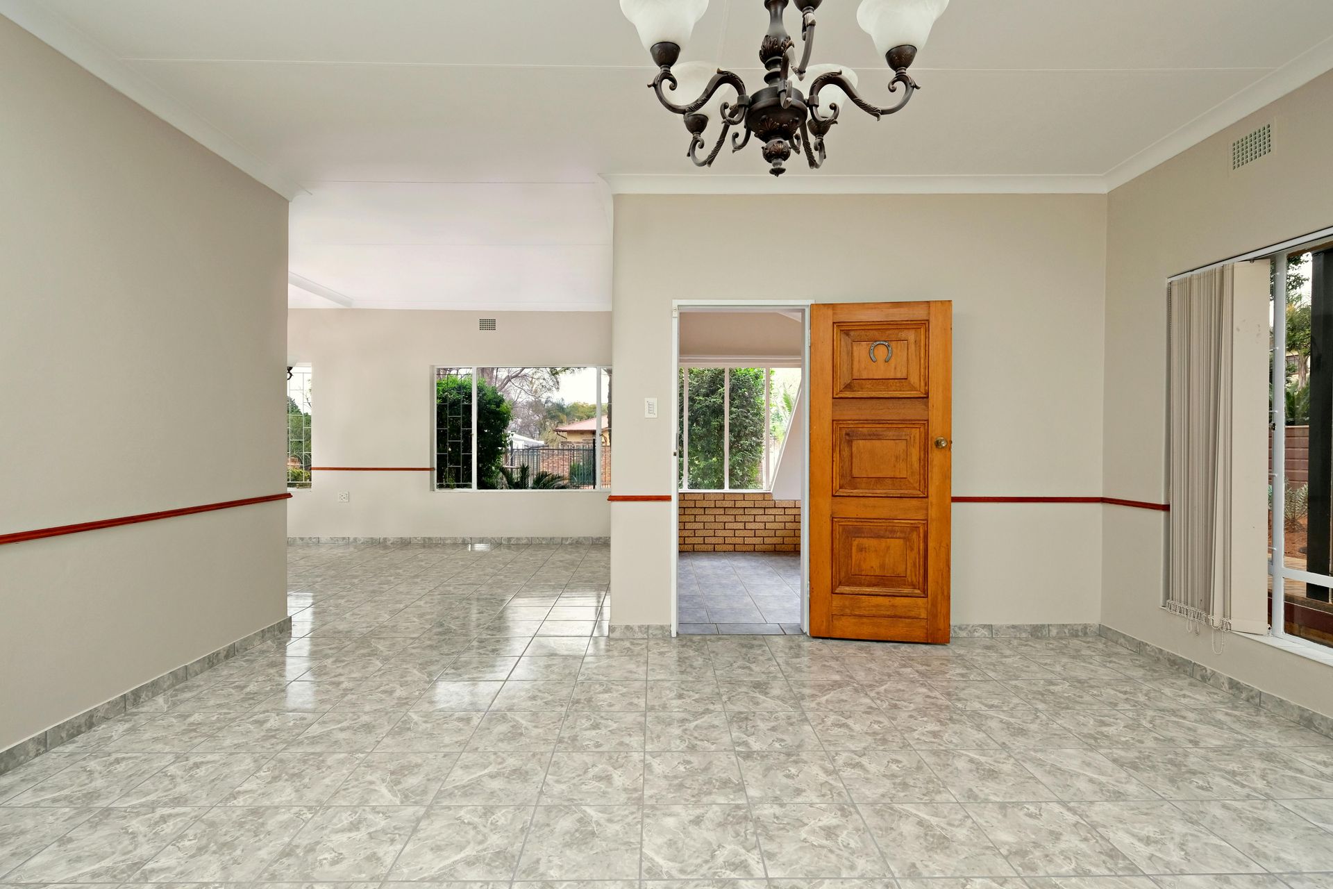 Empty tiled living room with beige walls, chandelier, and open wooden door to a small adjacent room.