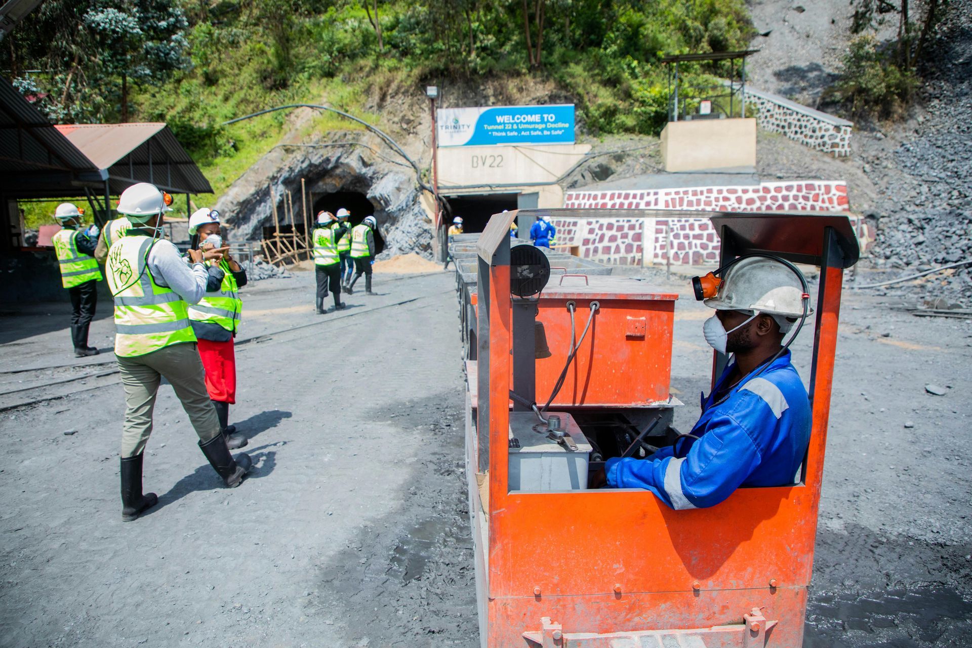 Road workers in safety vests and helmets on a mountain road, with a worker seated in an orange utility cart