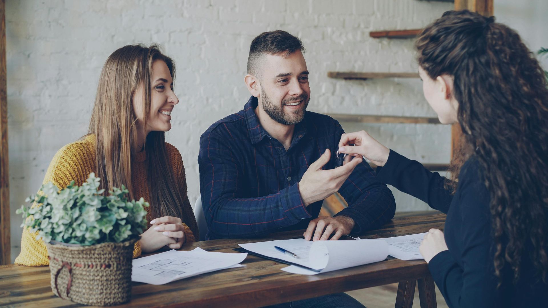 Three people in a meeting, smiling and discussing documents at a wooden table.