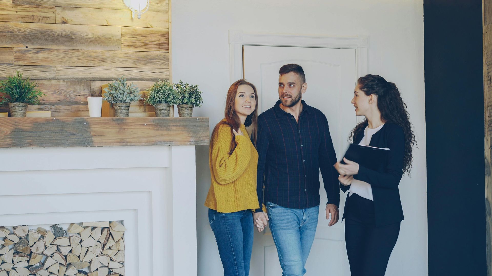 Three people standing and talking in a modern living room by a fireplace and wall art.