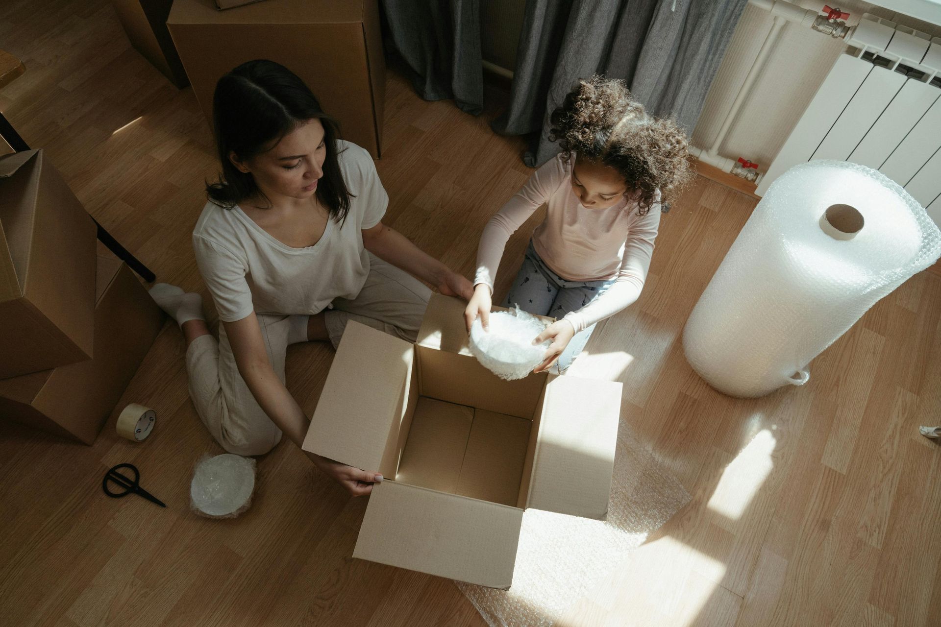 Two people packing items into cardboard boxes on a wooden floor beside a large white lamp.