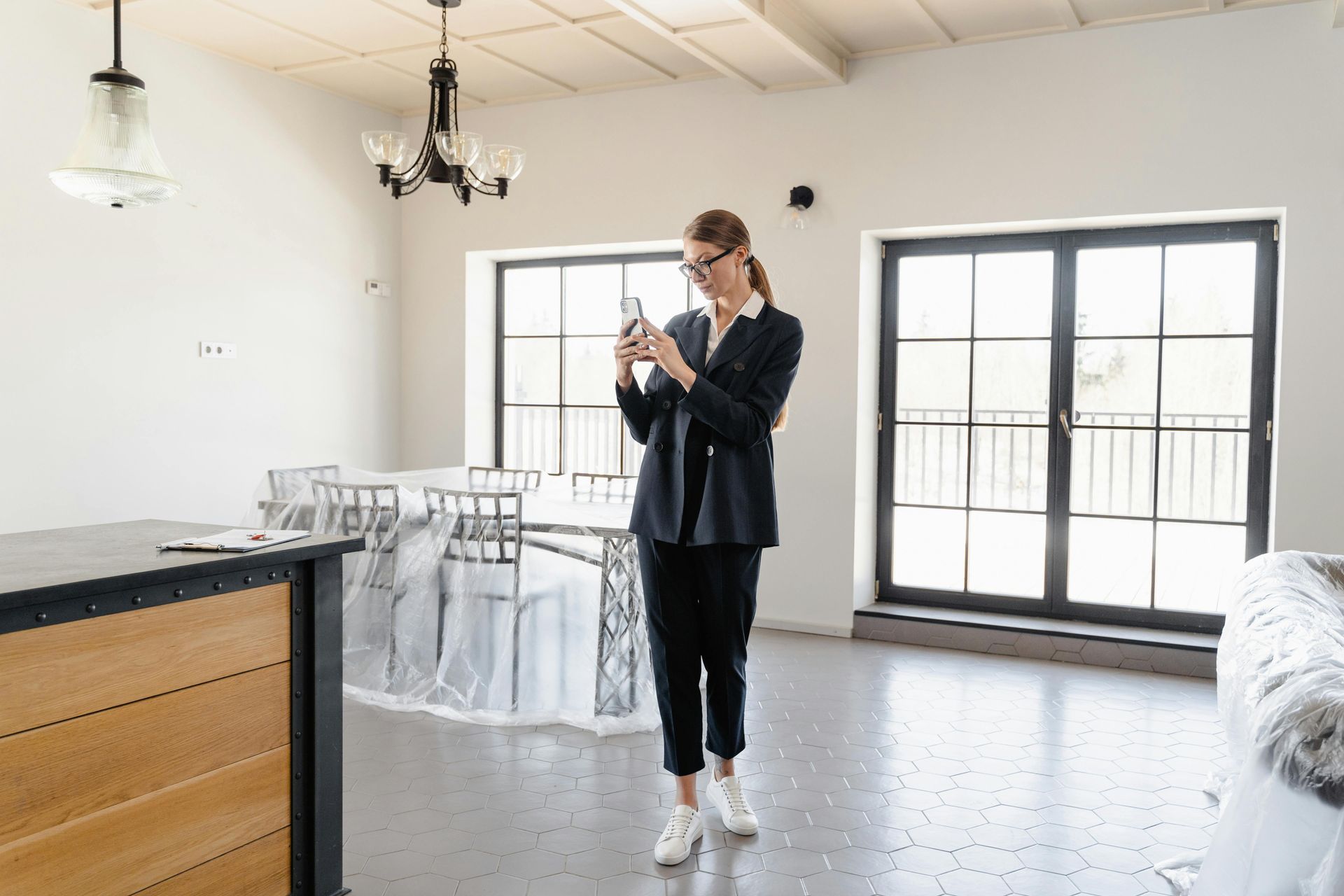Woman standing in a bright kitchen, looking at her phone near a dining area and large windows