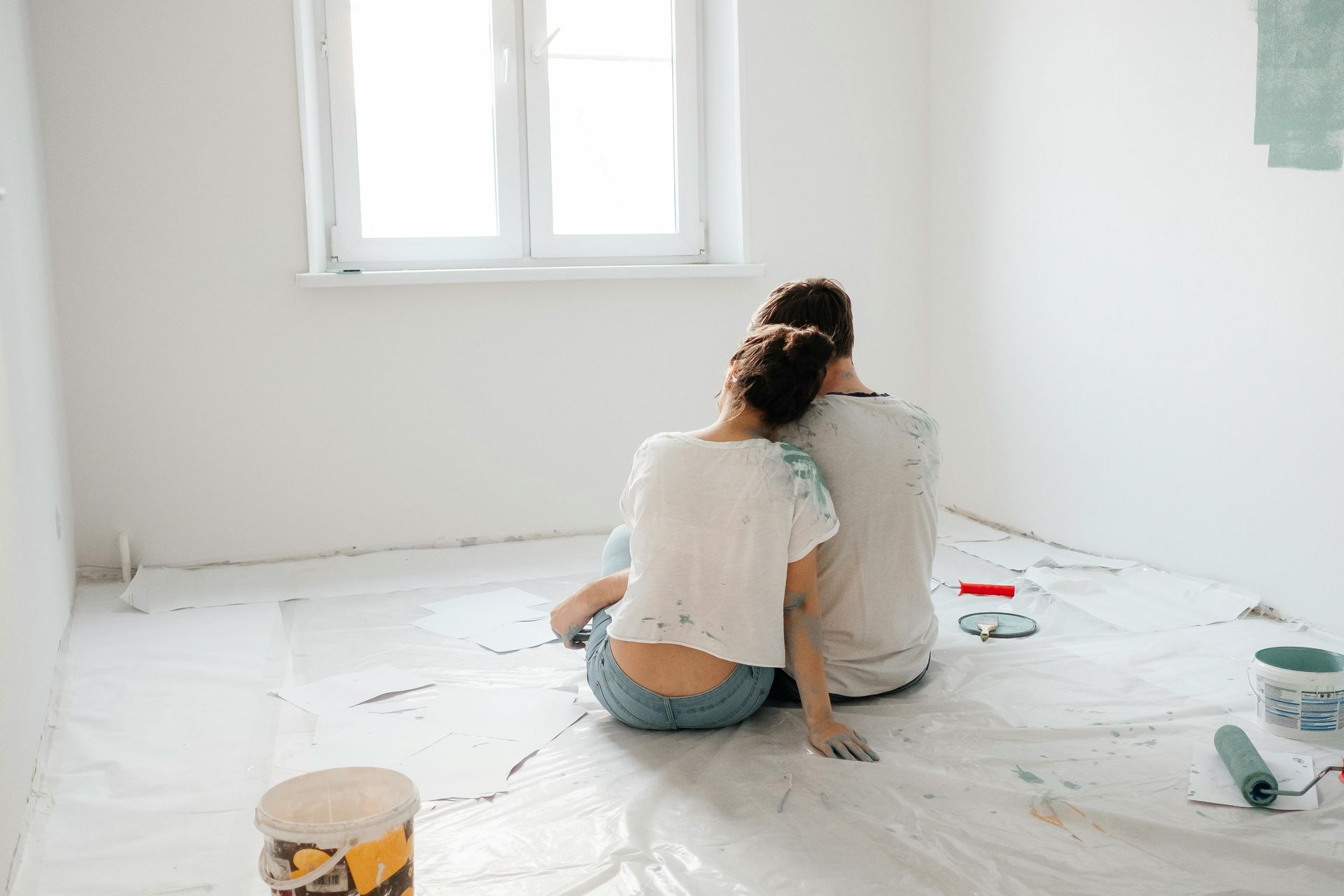 Two people sit on a floor covered in white plastic, painting a bright room with a window.