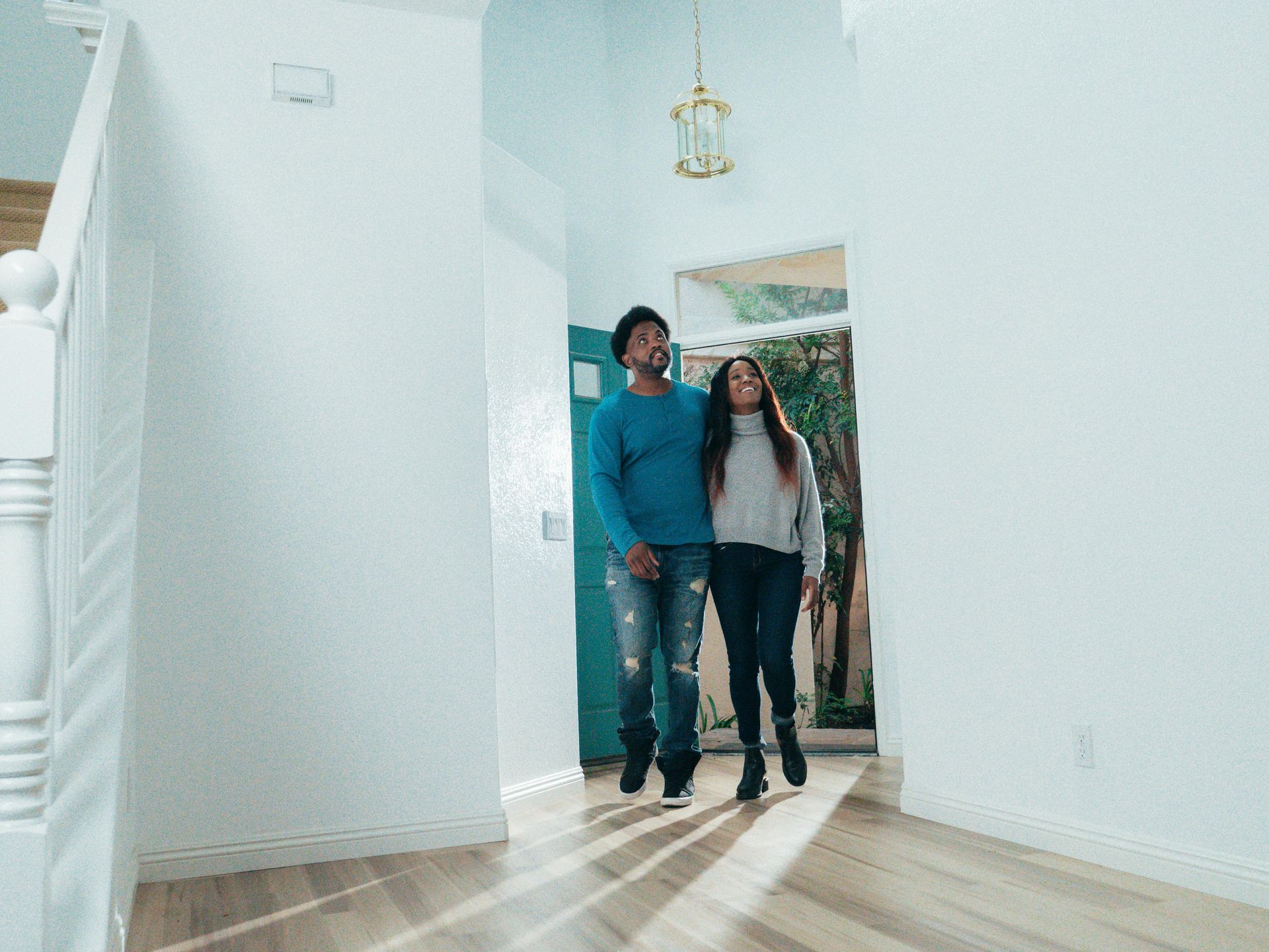Two people standing in a bright hallway near a doorway, with sunlight on the floor.