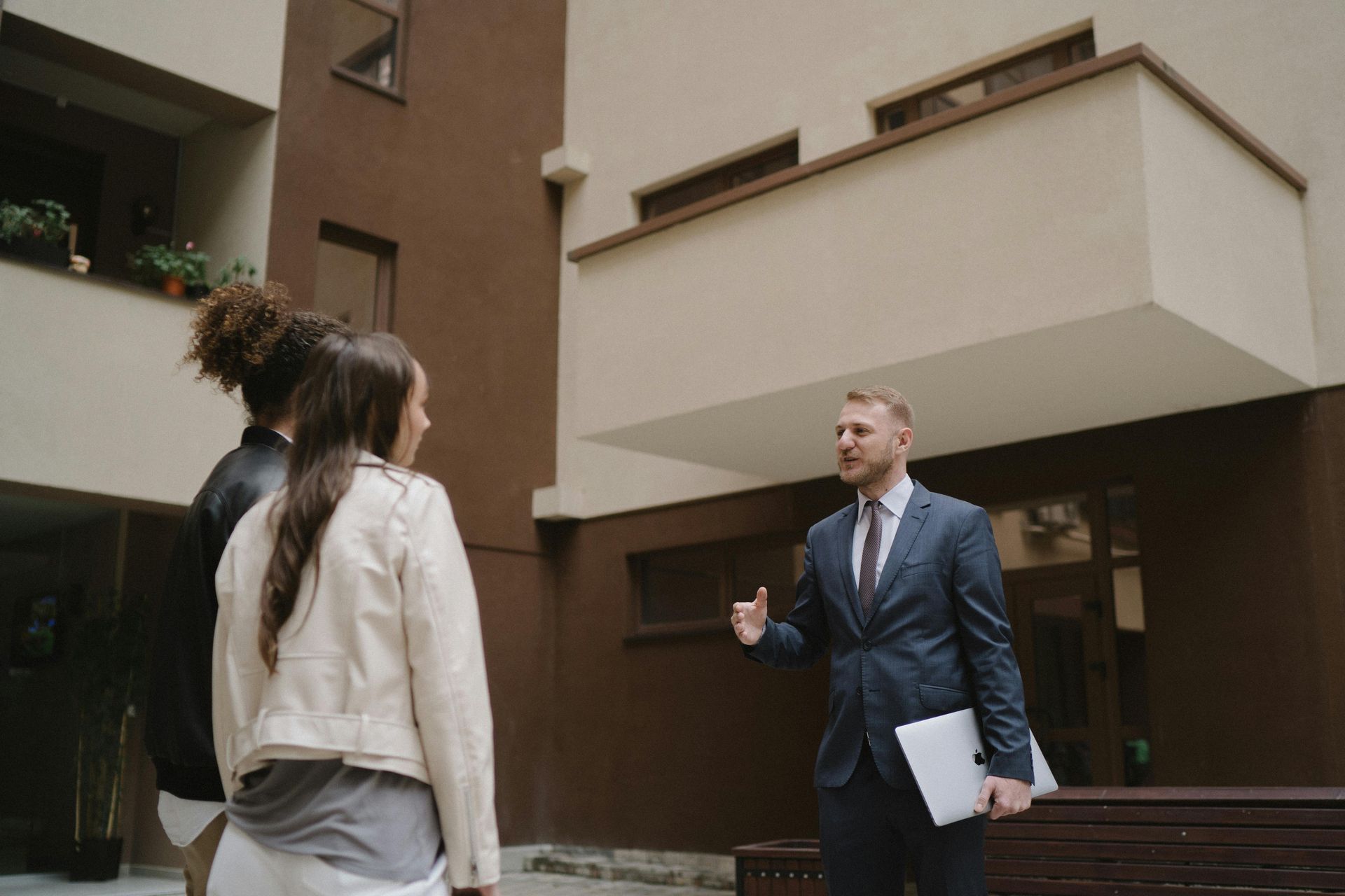 Three people talking in a courtyard; one man in a suit holds papers while gesturing.