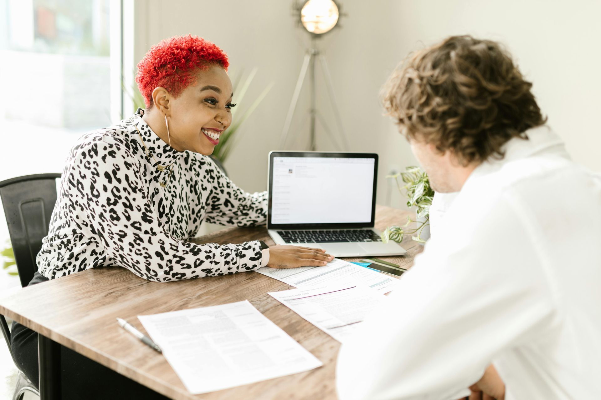 Two people in a bright office discuss documents across a wooden table with a laptop open.