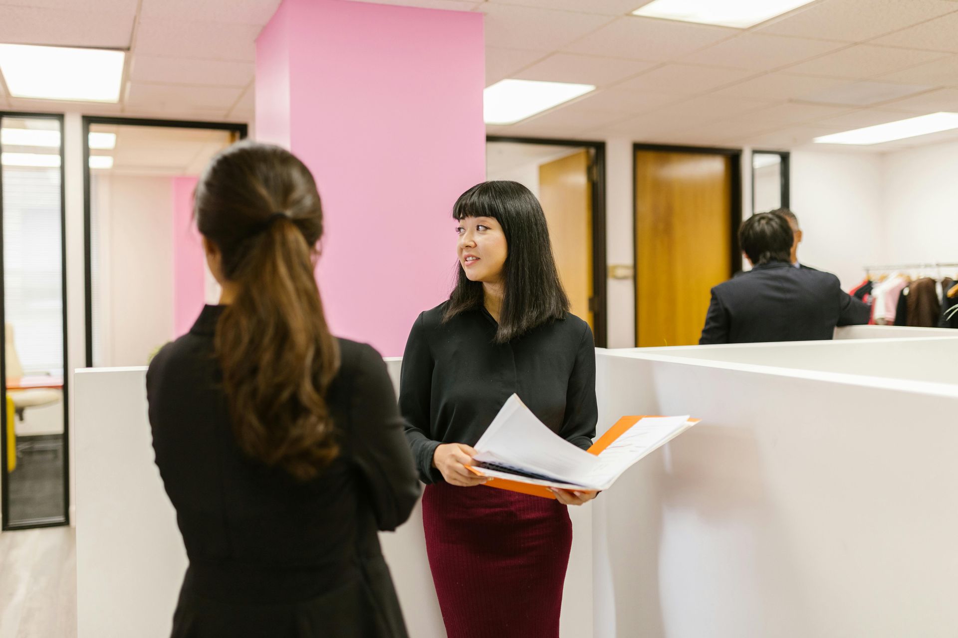 Two women talking in a bright office, one holding papers near pink and white cubicles.
