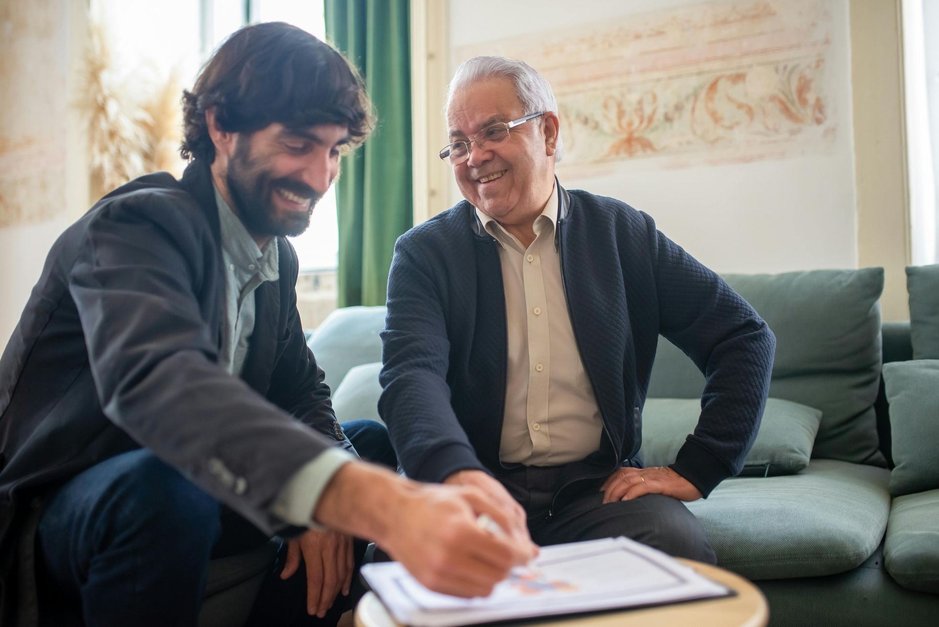 Two men smiling and reviewing a document together on a couch in a bright living room