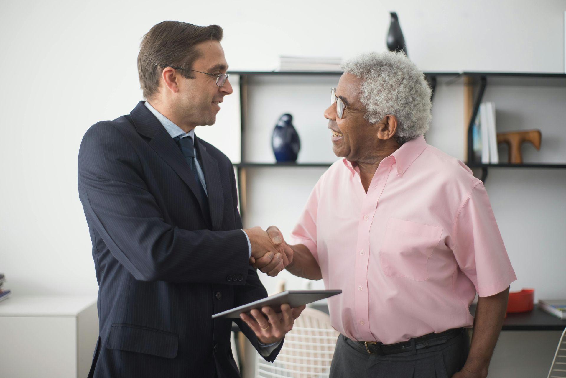 Two professionals shake hands in an office while one holds a tablet