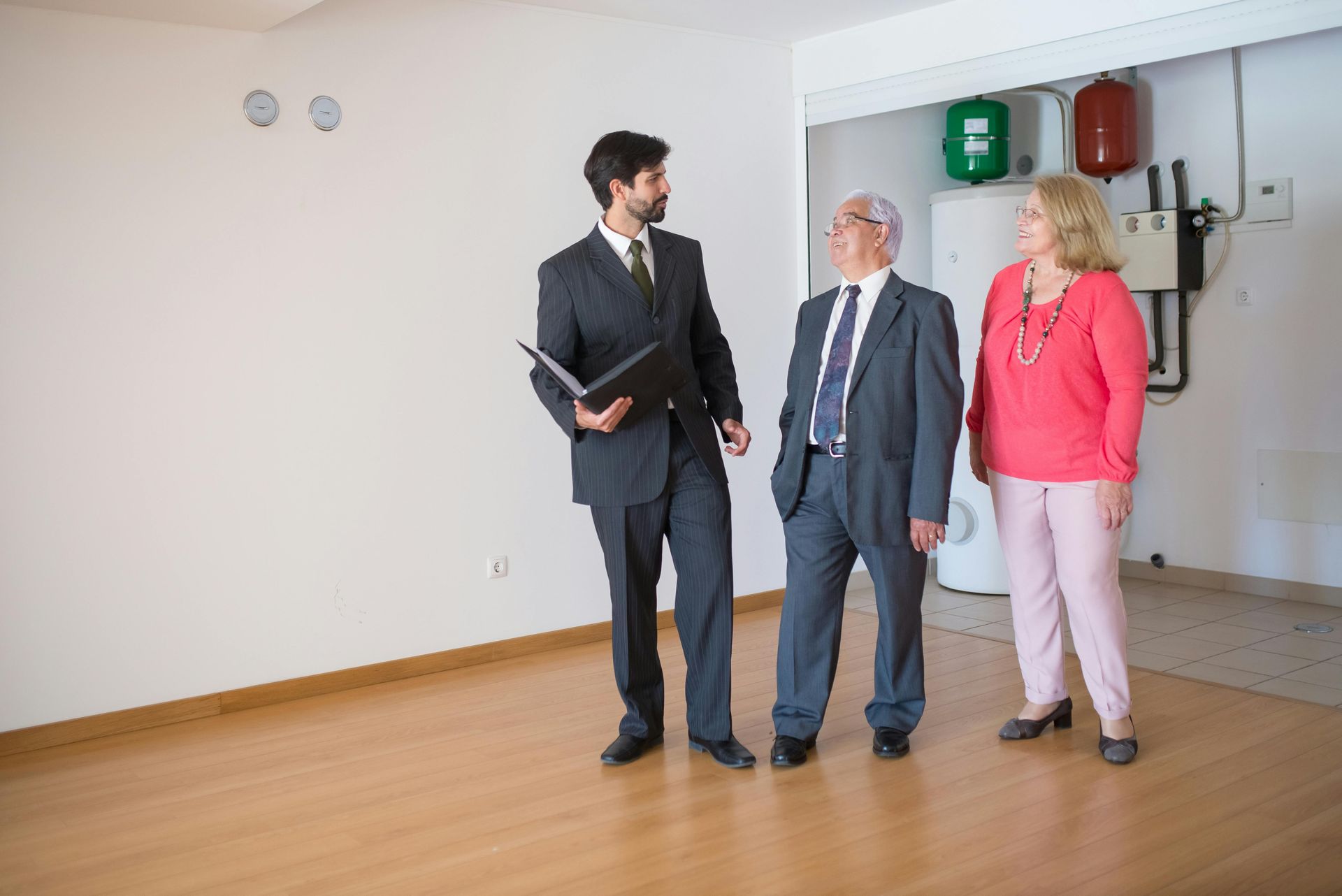 Three people in business attire stand and talk in a bright, empty room with wood floors.