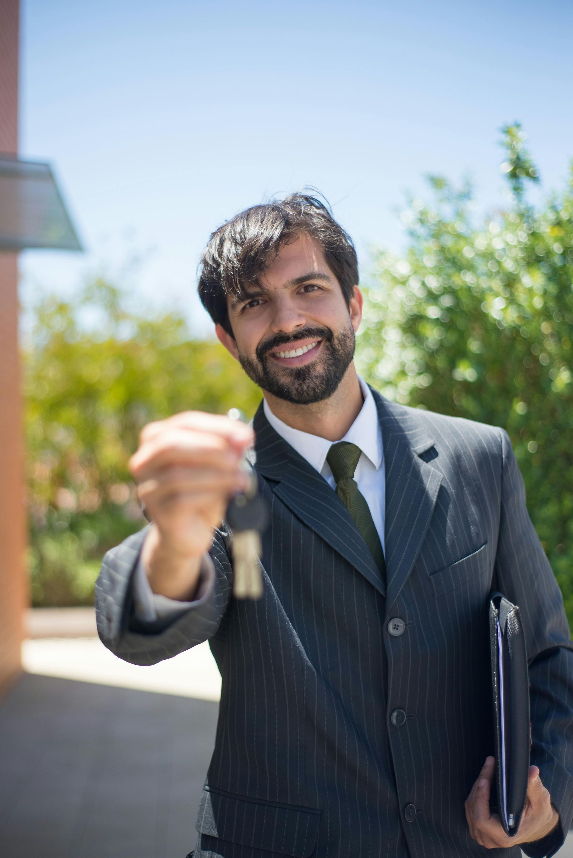 Smiling man in a suit holding out car keys and a folder outdoors on a sunny day