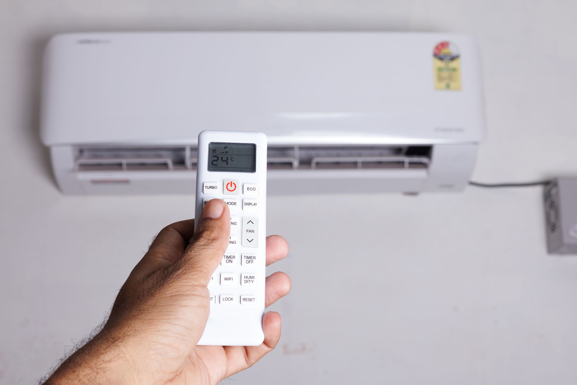A man's hand holding a remote control, raised and pointed at the air conditioning unit on the wall.