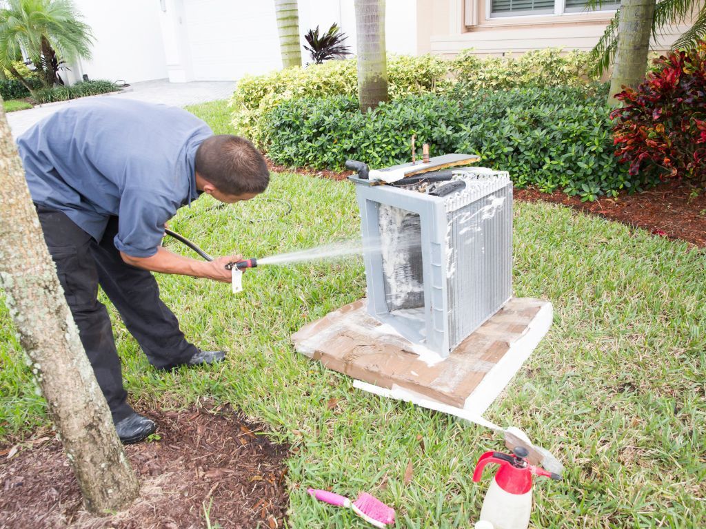 A man is spraying water on an air conditioner in the grass.