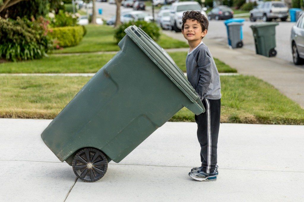 Boy pulling a green trash bin on a sidewalk in a residential neighborhood, smiling.
