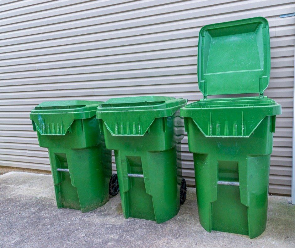 Three green trash cans, one lid open, lined up in front of a gray-striped wall.
