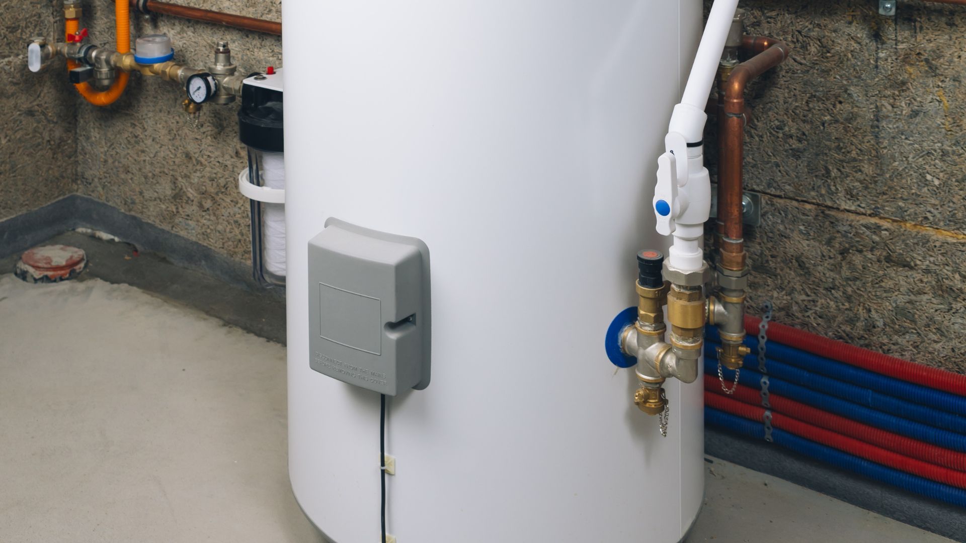 White cylindrical tank with attached gray box, pipes, and red and blue plumbing against a stone wall