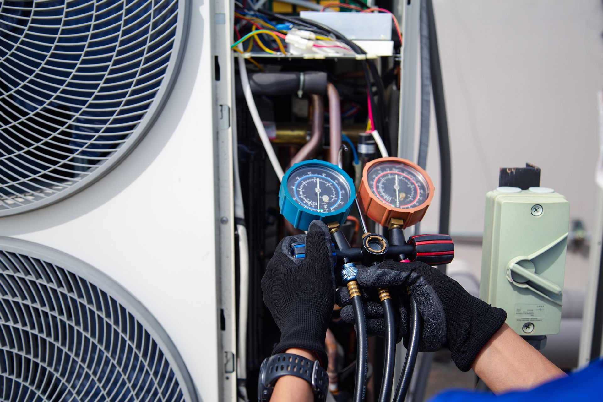 A technician wearing black gloves uses a manifold gauge set to service the open panel of an outdoor AC unit.