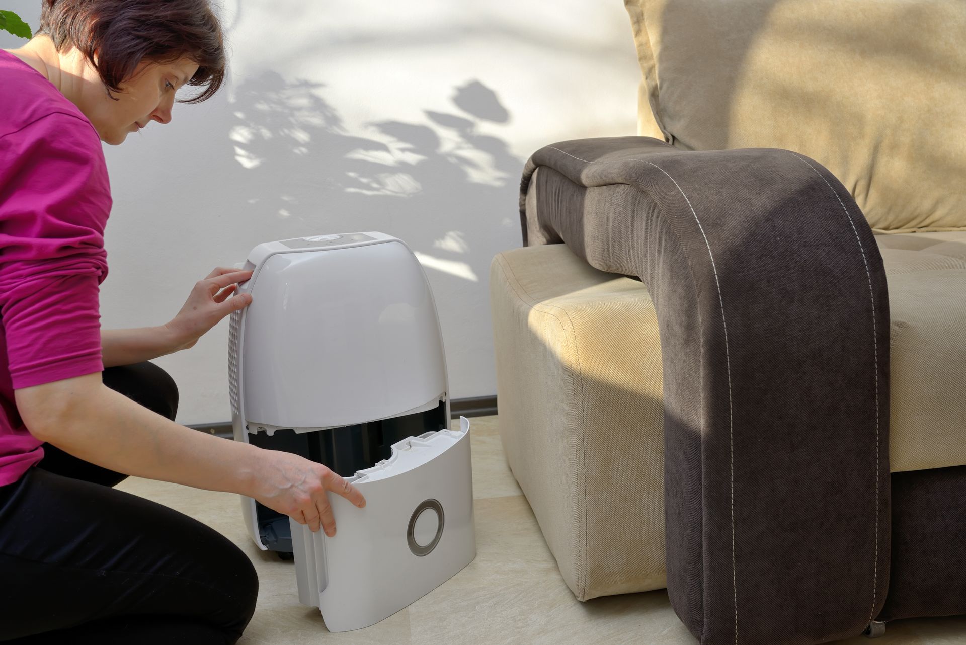 Woman kneeling next to a white humidifier, removing a component. Beside a beige and gray sofa.