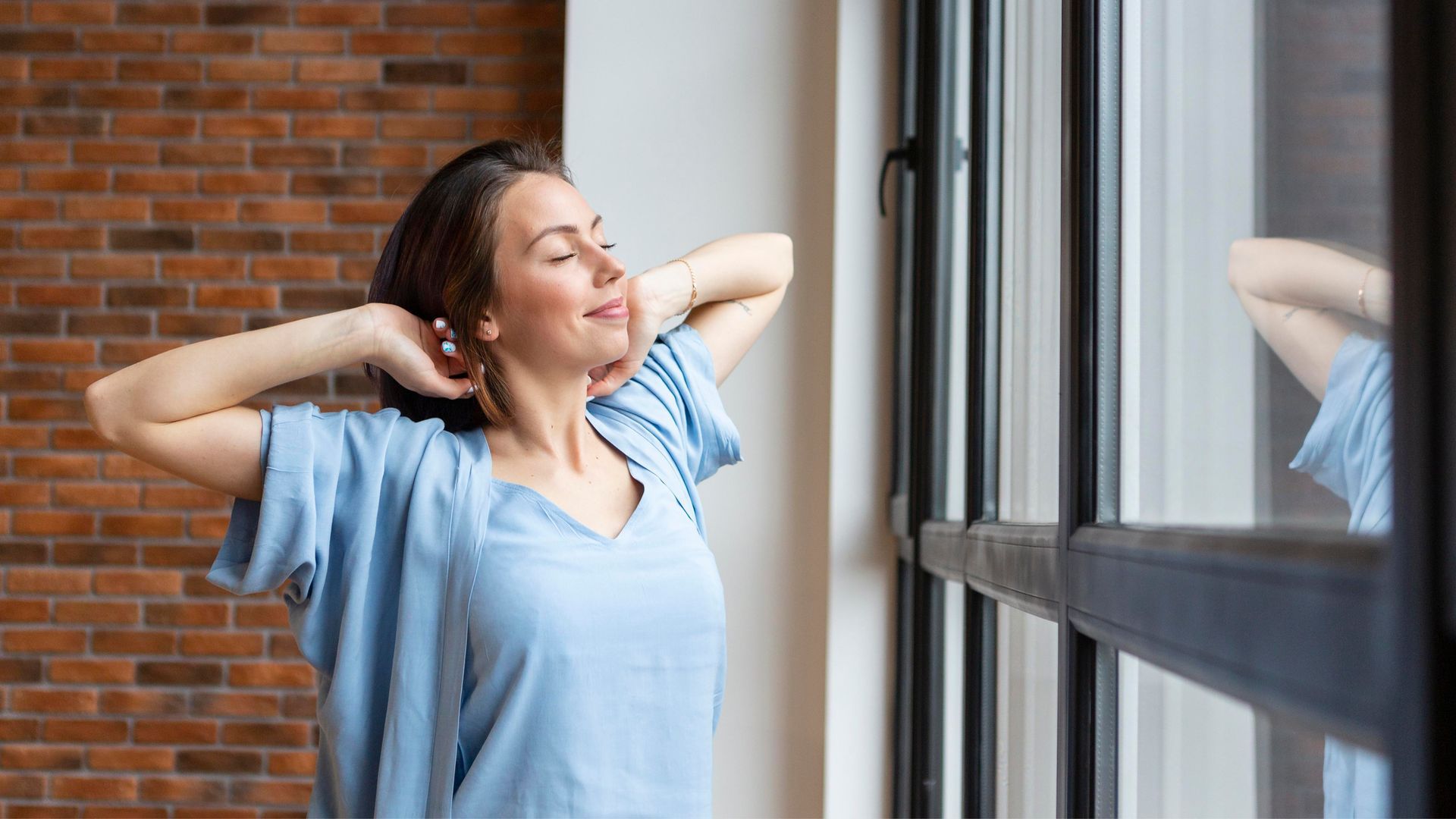 Woman stretching near a window with a brick wall, blue shirt, smiling.