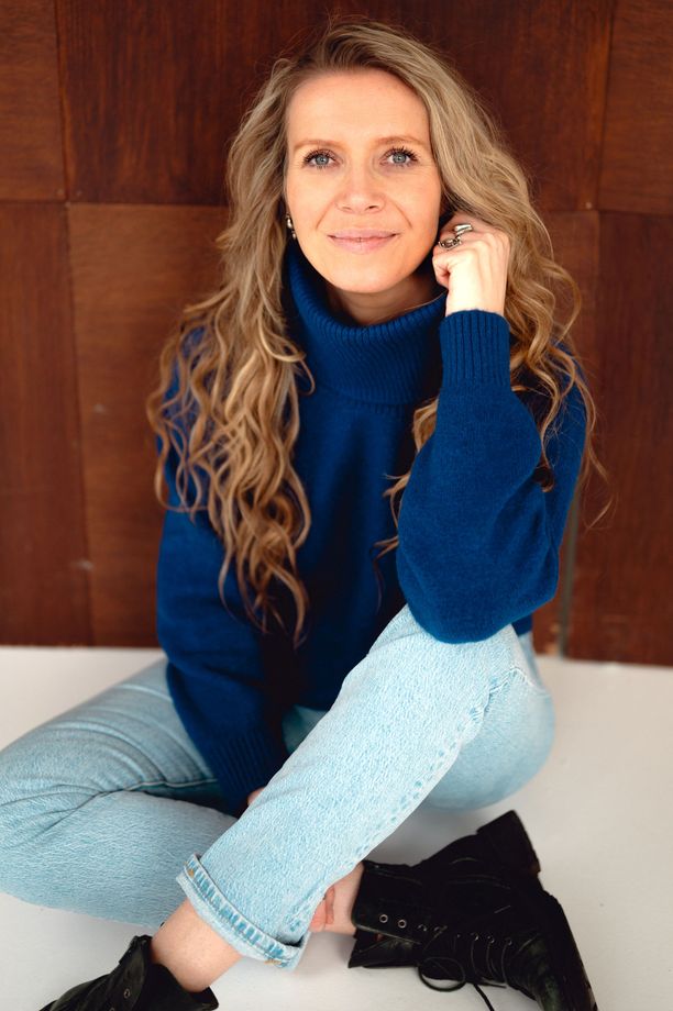 Debbie zit ontspannen tegen een bruine muur in een blauwe coltrui, spijkerbroek en zwarte laarzen.