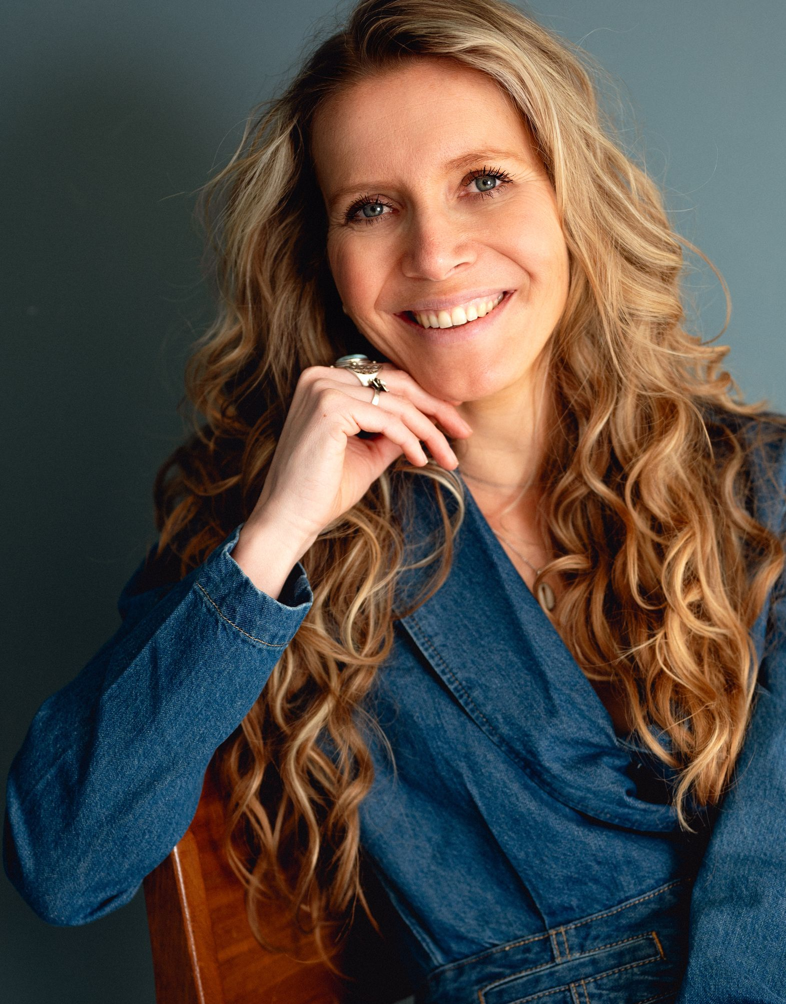 Debbie glimlacht met de hand bij haar kin, gekleed in een spijkerpak, tegen een blauwe achtergrond.