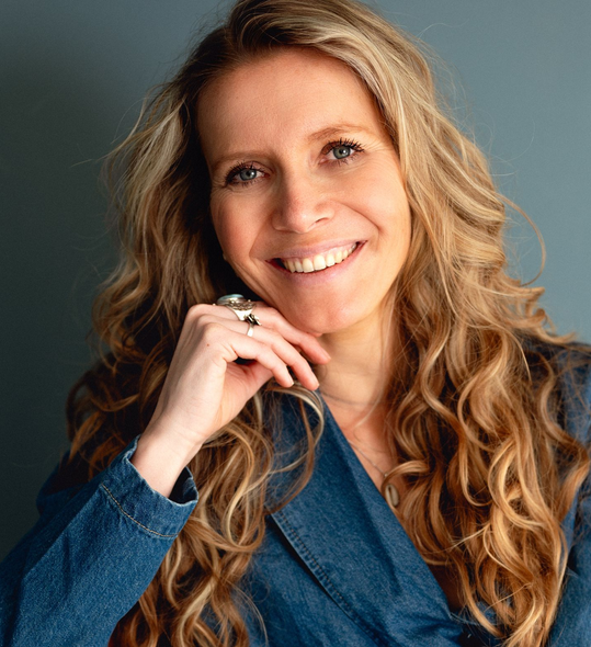 Debbie glimlacht met de hand bij haar kin, gekleed in een spijkerpak, tegen een blauwe achtergrond.