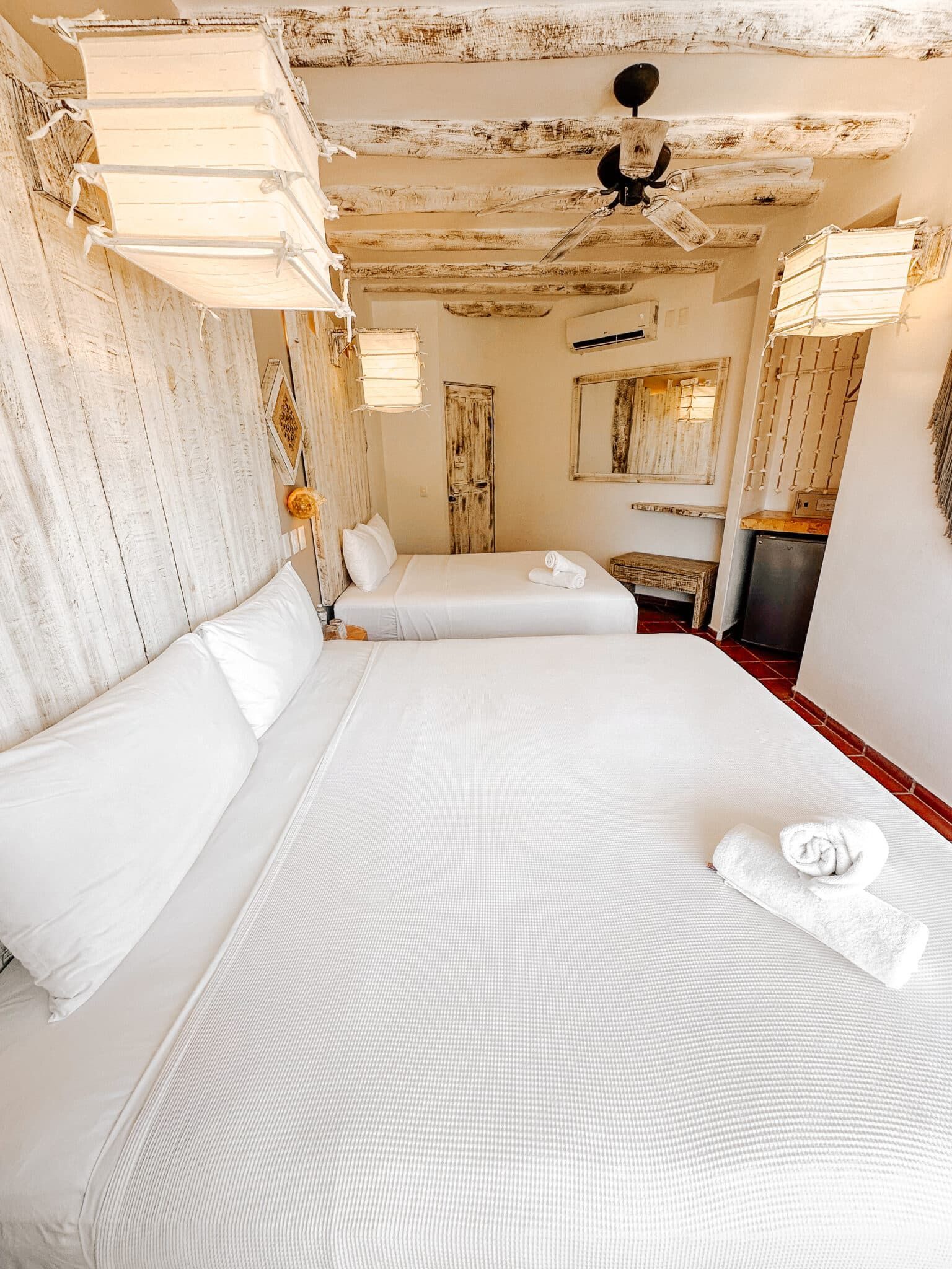 Dormitorio con dos camas, sábanas blancas, techo envejecido e iluminación decorativa.