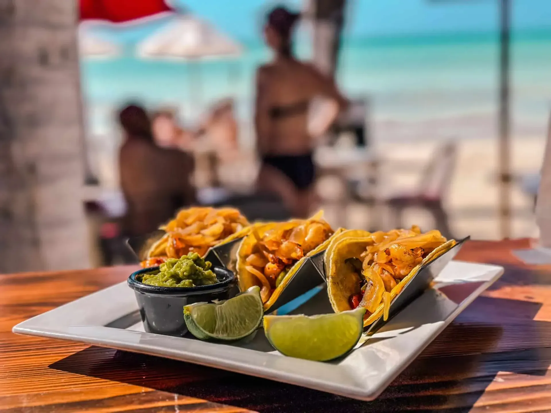 Tacos, guacamole y limón en un plato en un restaurante junto a la playa, con bañistas borrosos y el océano en el fondo.