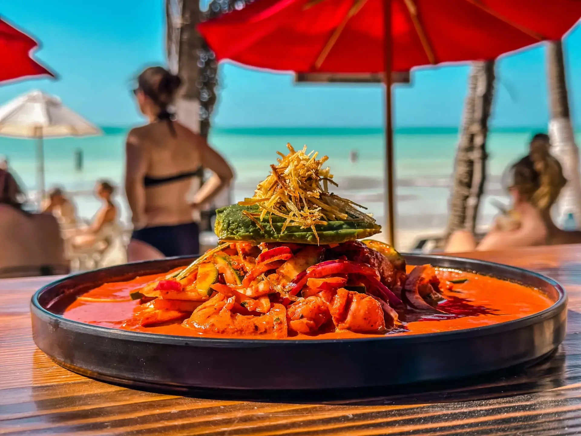 Plato de comida con mariscos en salsa roja, cubierto con aguacate y guarnición frita; escena de playa de fondo.