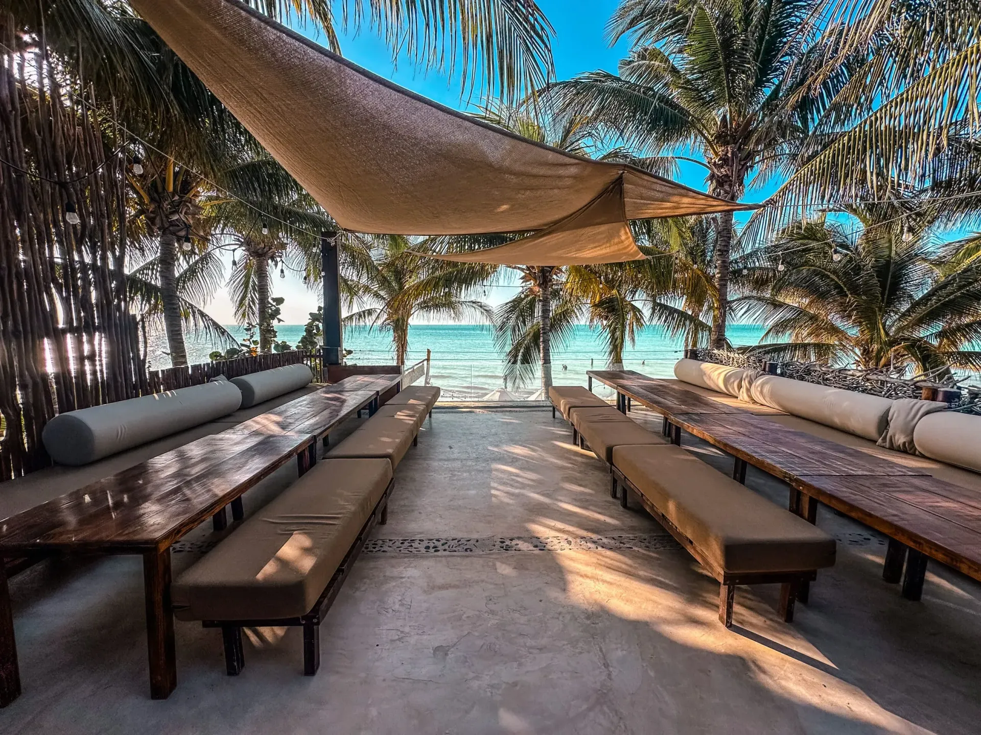 Restaurante de playa al aire libre con asientos a la sombra, vista al mar y palmeras.