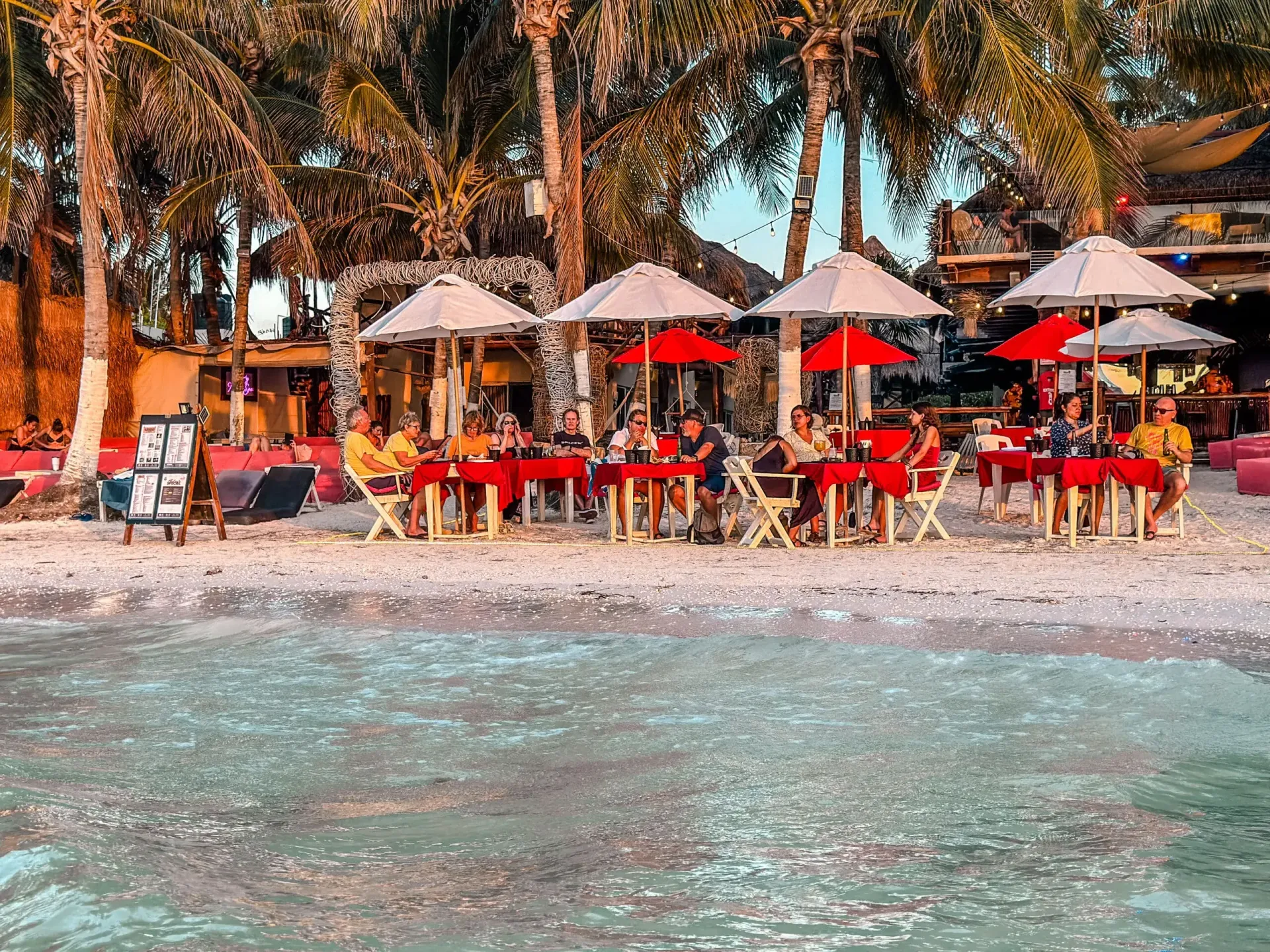 Restaurante junto a la playa con mesas y sombrillas cerca del agua. Palmeras y gente cenando.