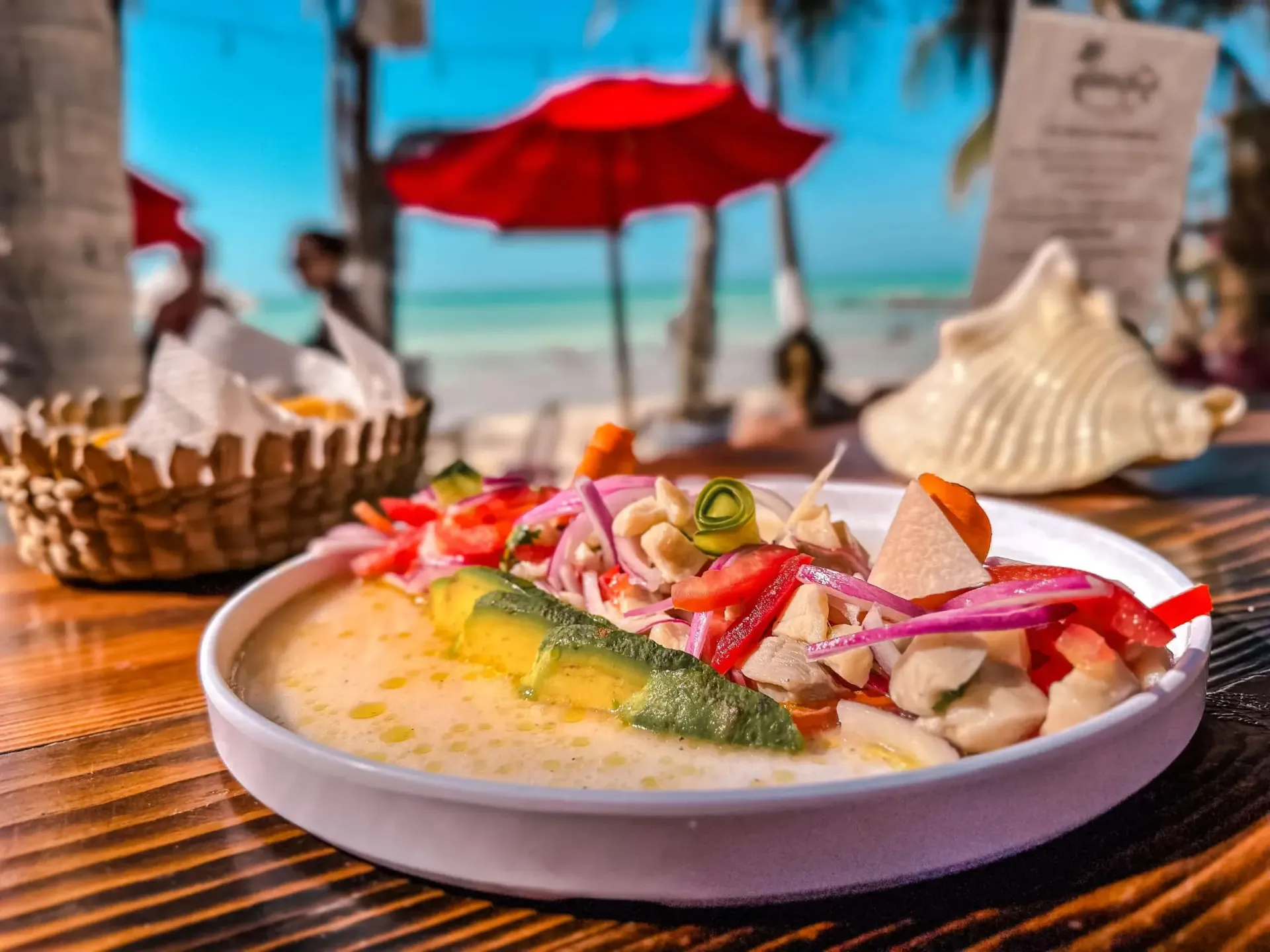 Plato de ceviche con rodaja de aguacate en una mesa cerca de una playa, sombrilla roja en el fondo.