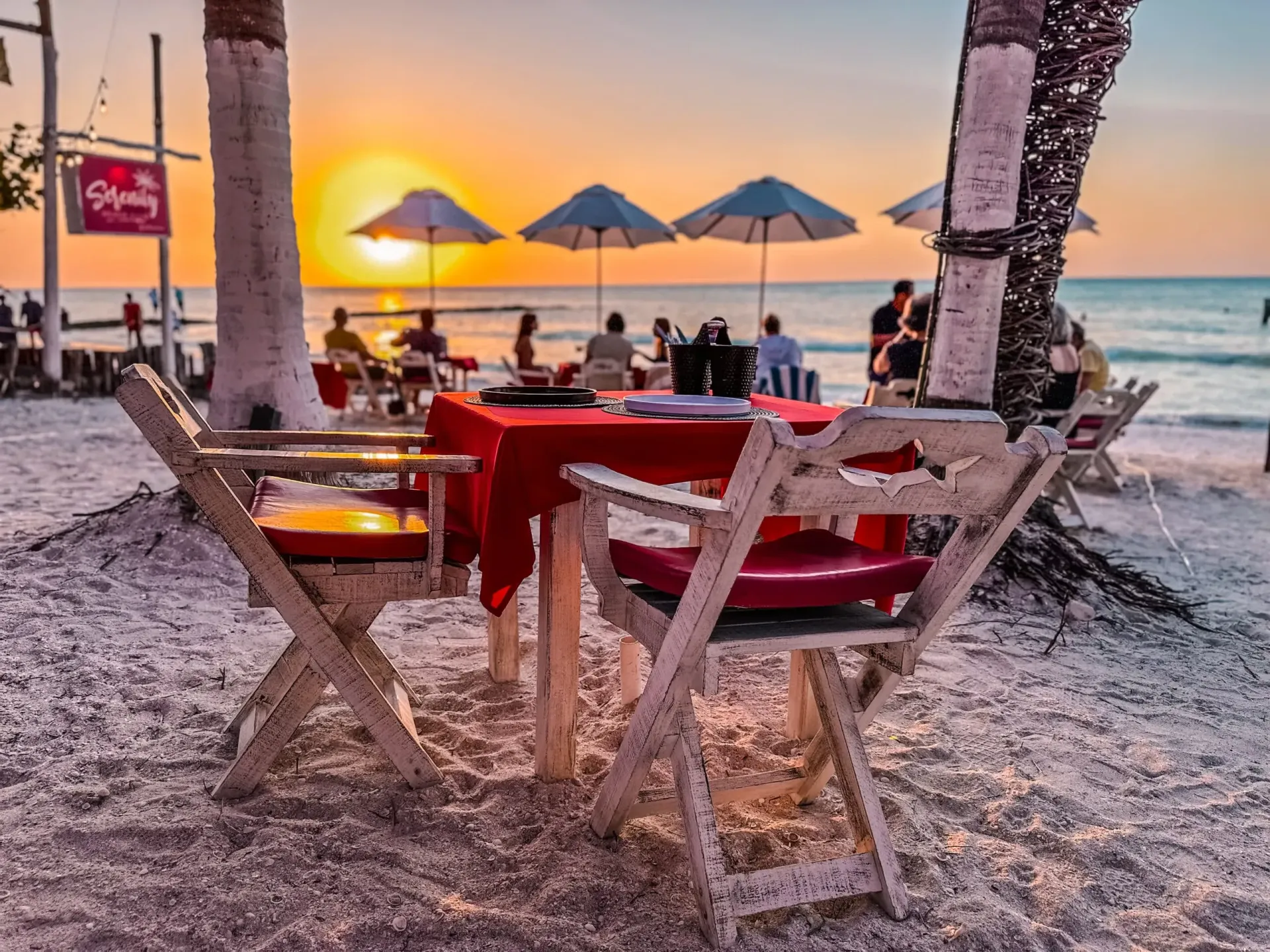 Mesa frente a la playa para cenar al atardecer; mantel rojo, sillas blancas.