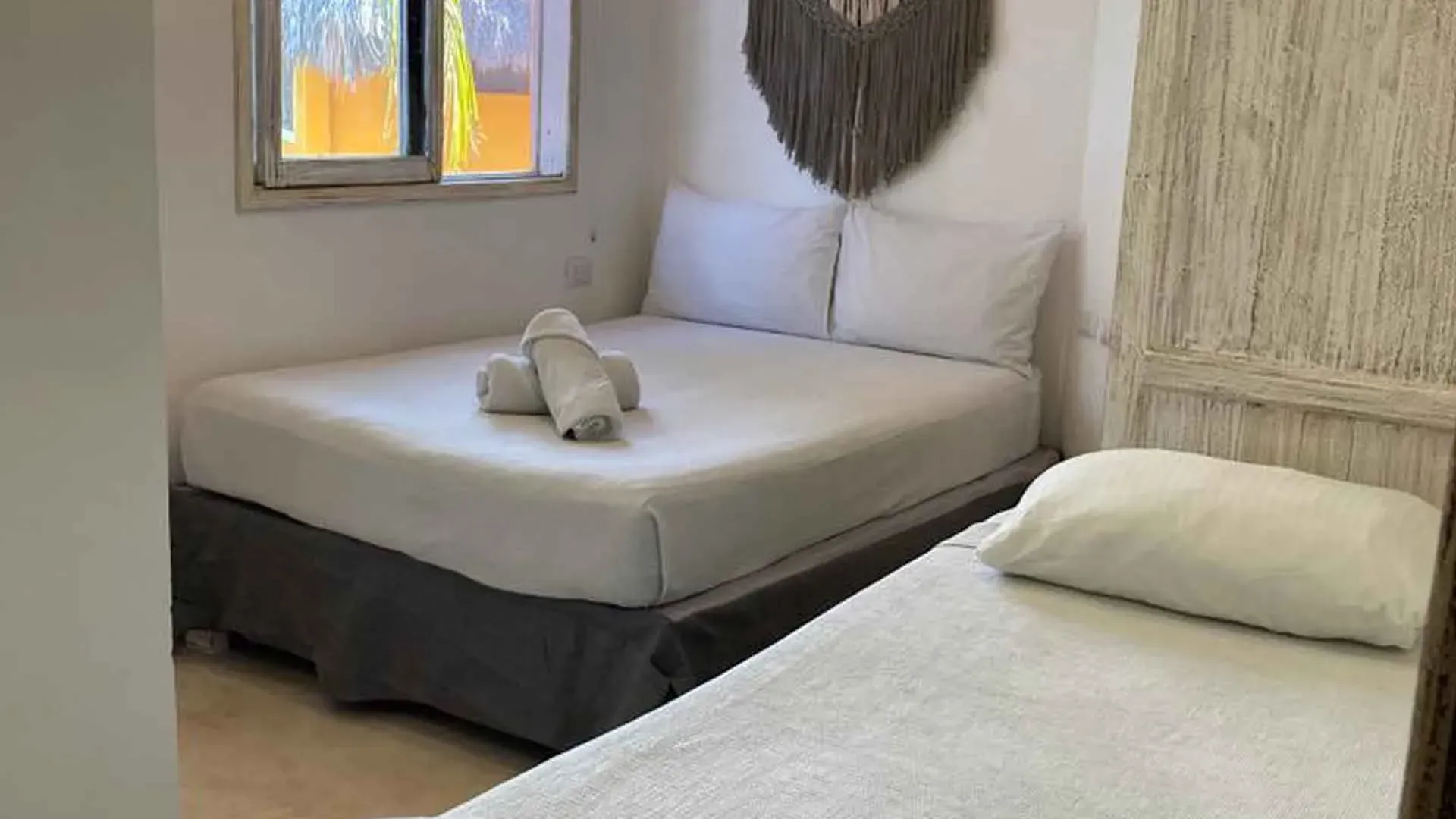 Dormitorio con dos camas; ropa de cama blanca, faldón de cama gris, ventana pequeña, tapices decorativos en la pared.