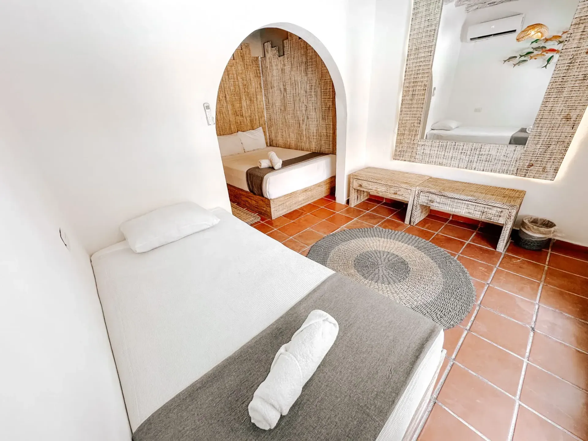 Interior del dormitorio: cama individual, manta gris, arco a otra cama, espejo, suelo de baldosas.